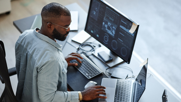 man working at a desk with two computer screens analyzing data and using technology for efficiency and productivity